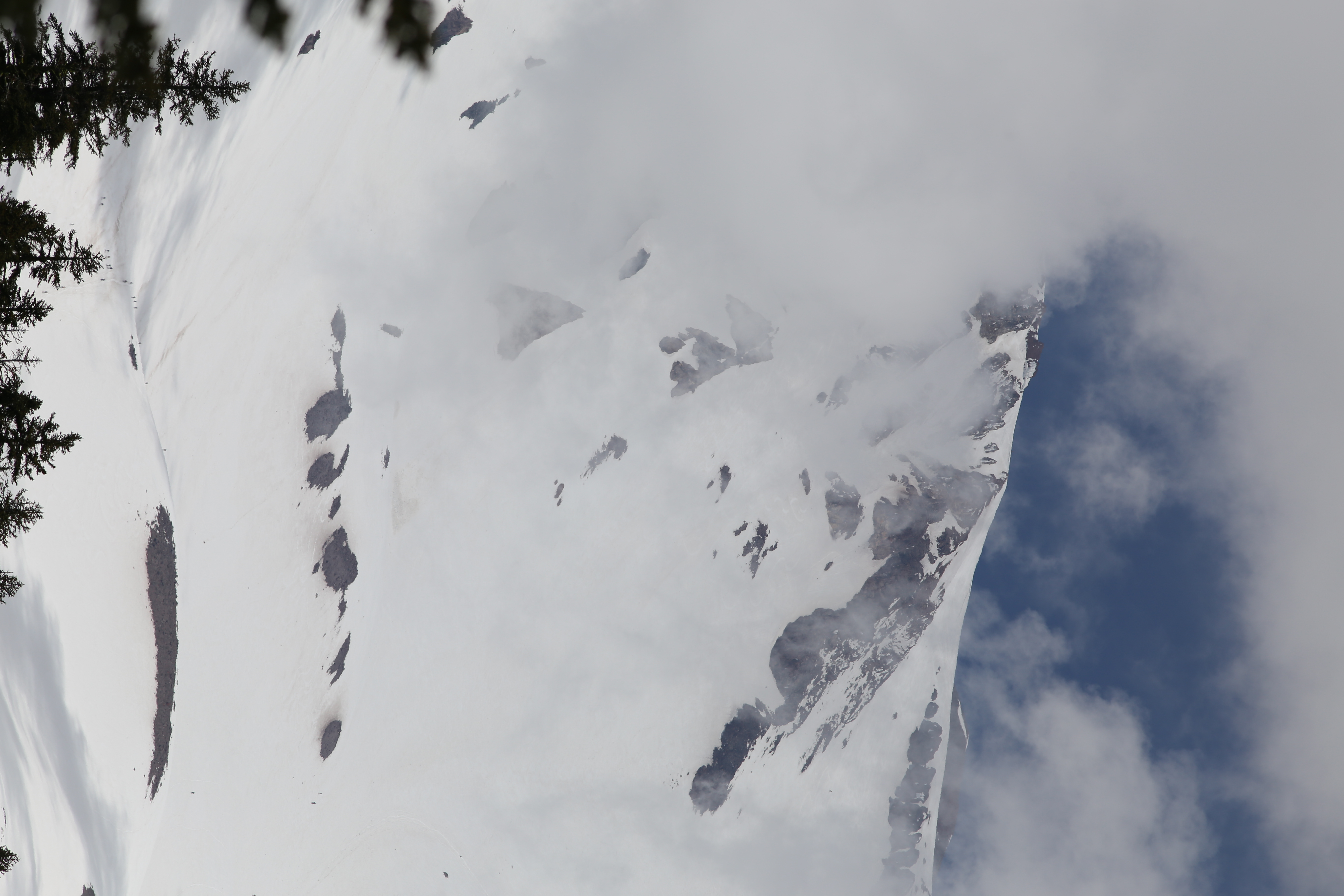 Close telephoto shot of Mt Shasta's steep snow-covered upper flanks with dark rocky outcroppings, clouds obscuring the summit and blue sky breaking through to the right