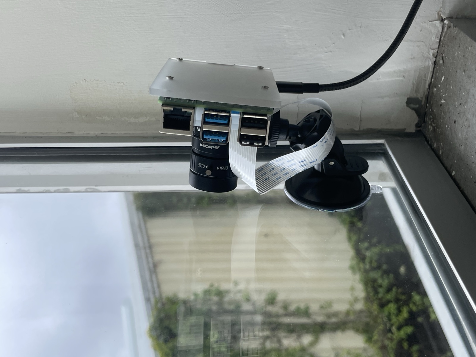 Raspberry Pi with an ArduCam lens, suction-mounted to a window frame overlooking a cloudy sky